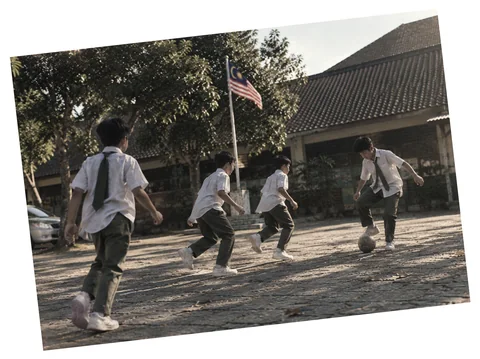 Murid sekolah bermain bola di halaman sekolah, dengan bendera Malaysia di belakang